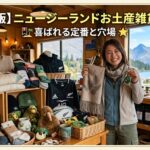 ニュージーランドのギフトショップで、マヌカハニーや羊毛製品、キウイのぬいぐるみなどのお土産雑貨に囲まれて笑顔で立つ女性。背景には美しい湖と山々が見える。